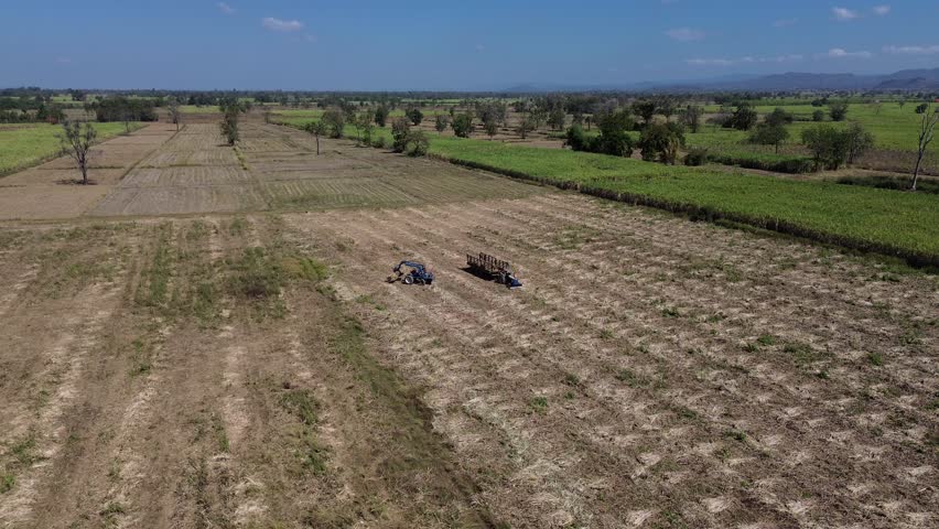 Aerial farmland tractor plowing field agriculture rural landscape harvest across cropland, dry plowed soil rows, green crop border and pasture, distant mountain range under blue sky, summer drought