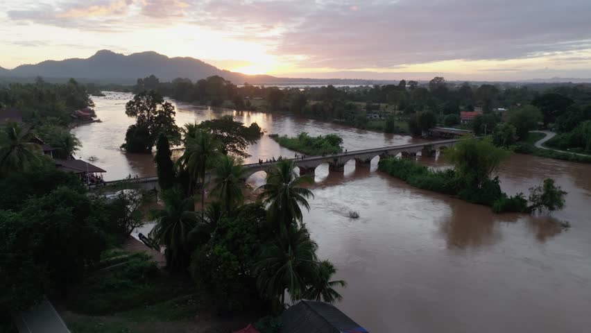 Aerial view of a stone bridge crossing a wide river at sunset in Don Det, Laos, surrounded by trees, palms, and village houses.