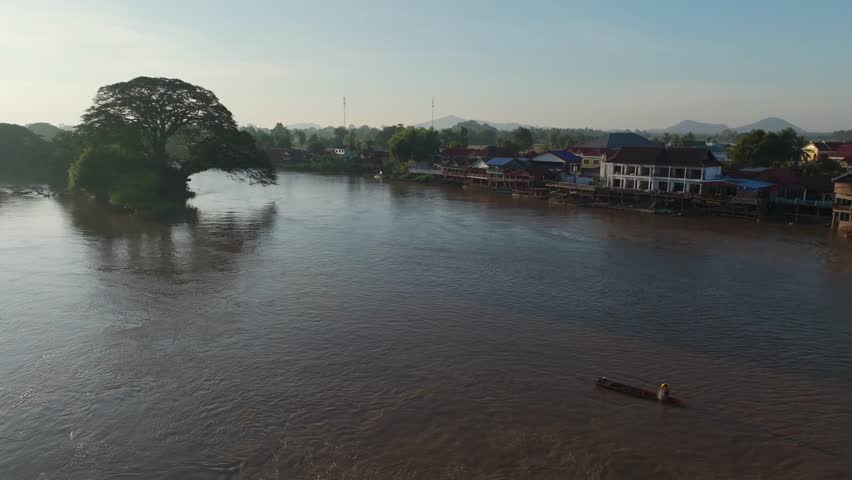 Small boat traveling across a wide river near Don Det, Laos, with riverside houses, trees, and calm evening light.
