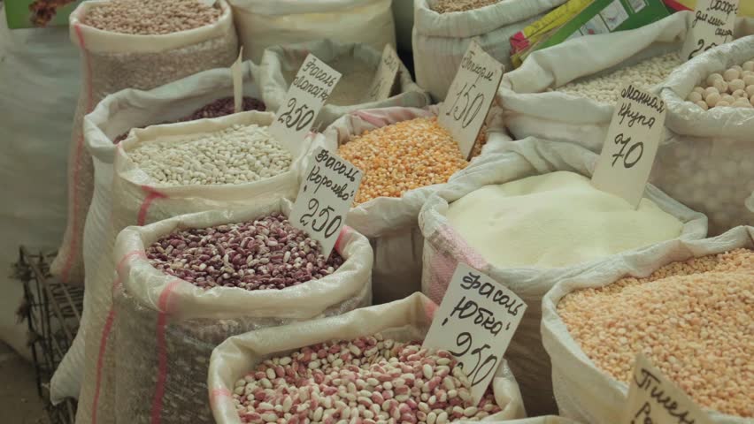 Shopper hand reaching into open sacks of beans, candid market stall scene with handwritten price tags, hand selecting pulses and corn, bulk food products and color contrast. Local bazaar trade