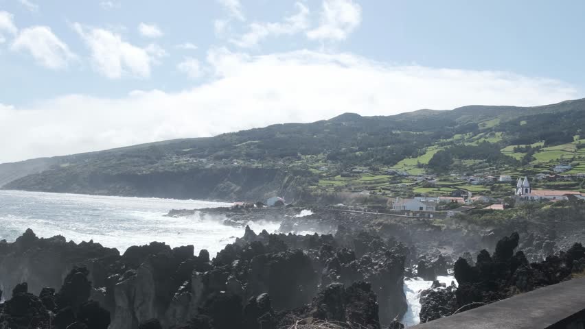 Atlantic waves crash against jagged lava rocks along the rugged shoreline of Pico Island in the Azores, with green hills and a small coastal village in the background.