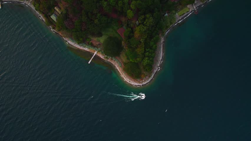 Zenithal aerial zoom in over Lake Atitlan shoreline showing forested land, pier and boat moving across deep blue water.