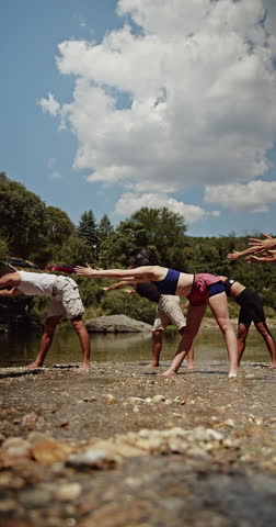 Friends enjoy a peaceful yoga session outdoors by a beautiful river. They stretch and find balance under the blue sky, promoting wellness and a healthy lifestyle in nature
