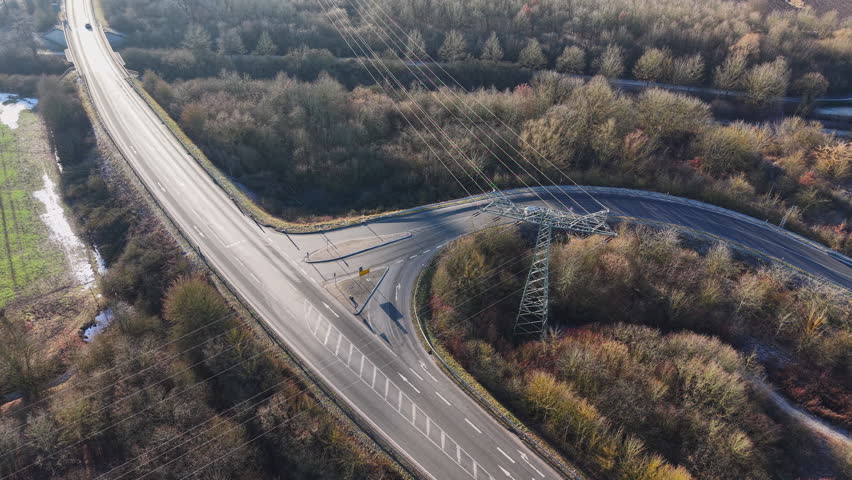 Aerial drone view of a road intersection surrounded by winter forest landscape, with high-voltage power lines crossing above the roadway. The scene shows modern infrastructure combining transportation