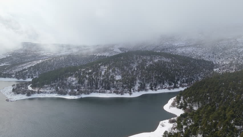 Aerial Shot of Snowy Forest Peninsula and Mountain Lake with Misty Hills in Winter Atmosphere