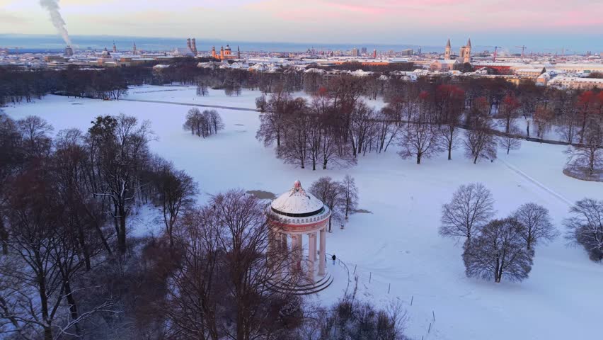 Luftaufnahme vom winterlichen Muenchen. Blick vom verschneiten Englischen Garten mit Monopteros auf die historische Altstadt und das Stadtzentrum bei einem wunderschoenen Sonnenaufgang