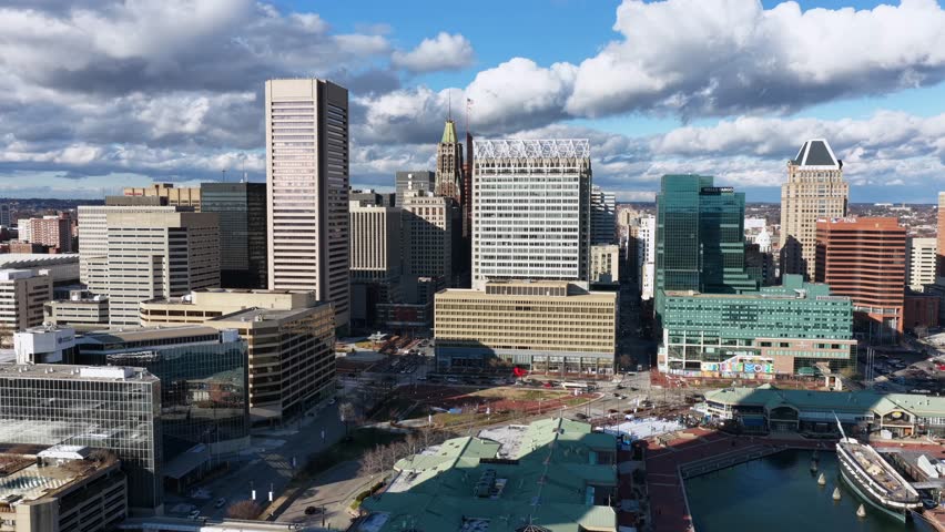 Aerial view of Baltimore’s Inner Harbor and downtown skyline, showing high-rise buildings, marina docks, waterfront attractions and dense urban neighborhoods under bright daytime sky.