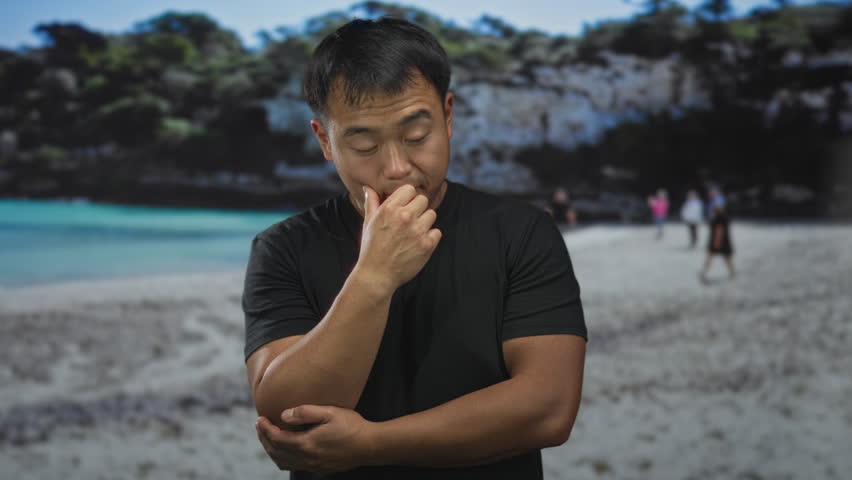 Man rubbing cheek and holding elbow on sunlit beach with turquoise water and massive rock cliffs overhead; fatigue.