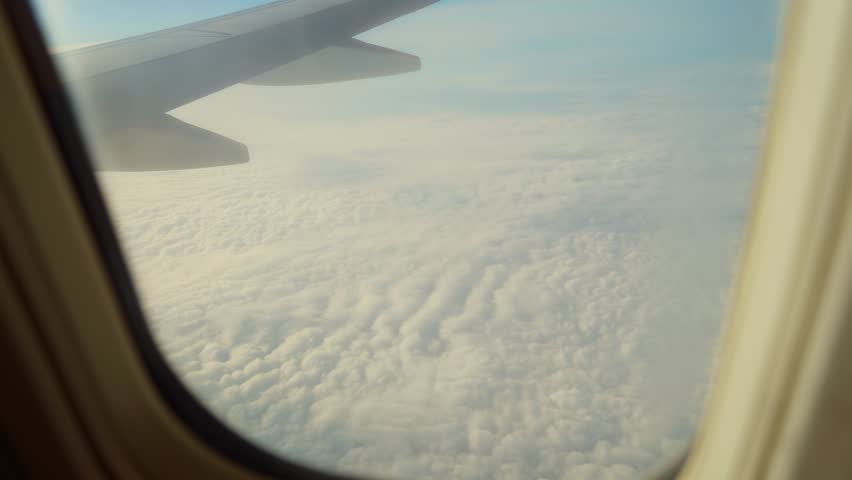 Jet flying above thick fluffy clouds in soft sunlight, view from window porthole. Plane cruising through clear blue sky over dense white cloud layer. Aircraft soaring high above textured cloudscape