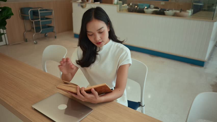 High-angle view of a young asian woman sitting at a table and reading a book. A closed laptop is next to her in a modern coffee shop