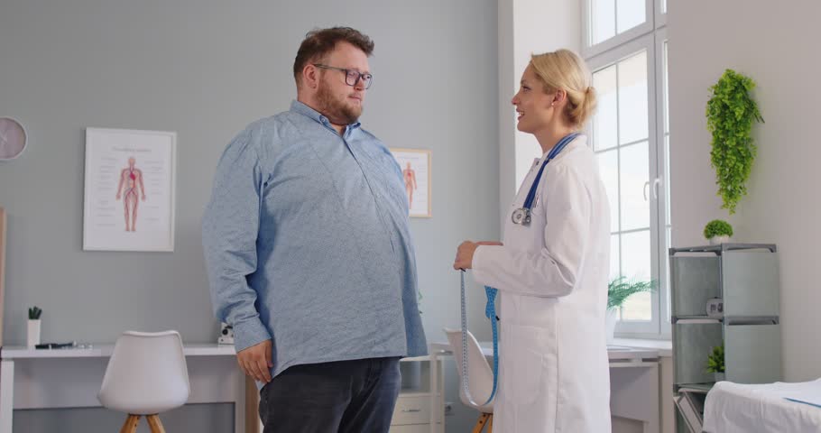 Clinician helps an overweight fat patient in a clinic exam, using tape to record waist measurement and provide obesity counseling on diet and health risks. Nutritionist measures patient waist