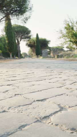 Young man and woman walking on the cobblestones of the Appia Antica, a famous ancient Roman road surrounded by pine trees and historical ruins, enjoying a sunny day of sightseeing in Rome, Italy