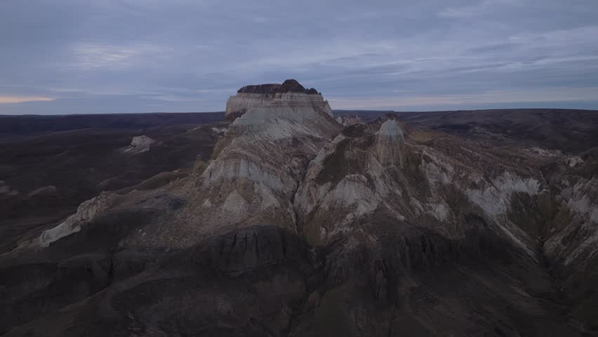 Aerial view of a rugged mountain range with layered rock formations, showcasing earthy tones under a cloudy sky, Shetpe, Mangystau Region, Kazakhstan.