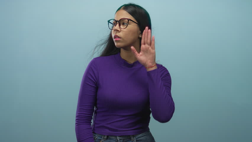 Young latin woman holds hand to ear listening in blue studio; curiosity discovery awareness exploration.