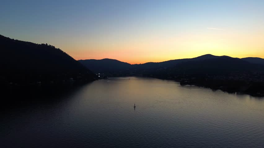 Aerial view of the serene Lake Como water reflecting the vivid sunset colors, framed by dark, silhouetted mountains, creating a tranquil scene, Lombardy, Italy.