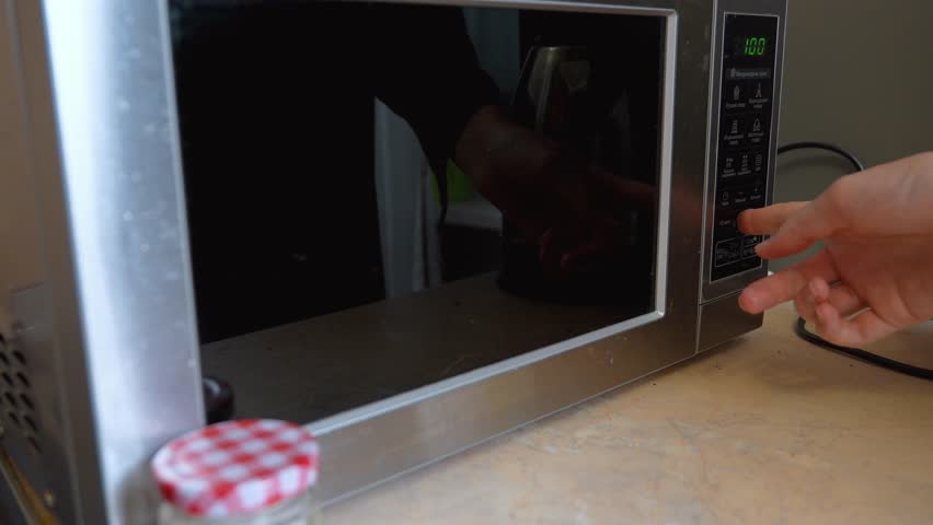 Close up view of a person's hand placing a glass inside a modern stainless steel microwave oven, closing the door and starting the timer to heat up the contents on a kitchen counter