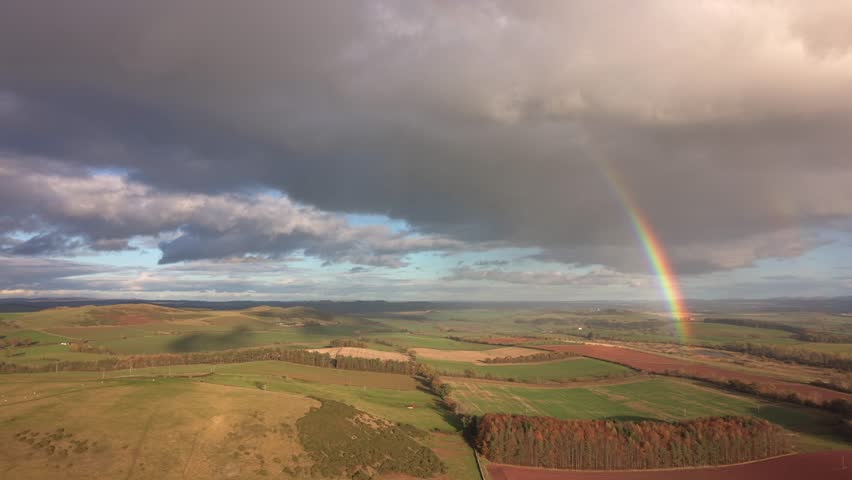Aerial view of a picturesque landscape featuring a vibrant rainbow arcing across fields and hills under a dramatic sky, Melrose, Scotland, United Kingdom.