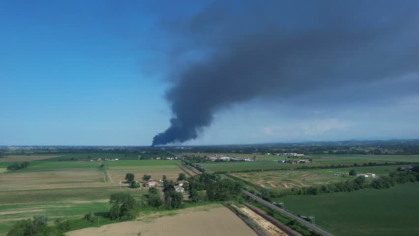 A large plume of dark smoke billows from an industrial site, spreading across cultivated farmland under a clear blue sky, indicating severe air pollution