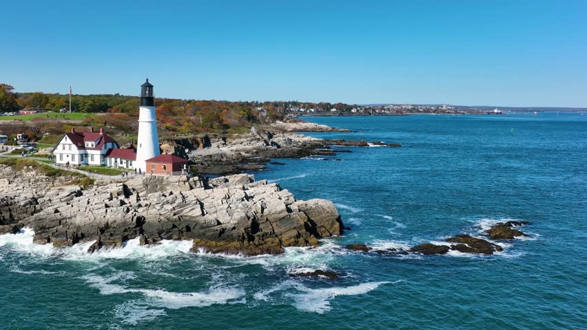 Aerial view of Portland Head Light with white waves crashing against the rocky coast under clear blue skies, Cape Elizabeth, Maine, United States.