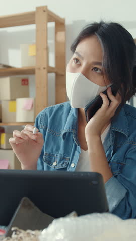 Vertical Screen : Young Asia businesswomen wear face mask using mobile phone call receiving purchase order and check product on stock work at home office. Small business owner, online market delivery.