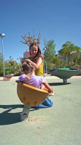 Young girl laughing and having fun with her smiling mother on a playground merry go round. Happy family enjoying a sunny day outdoors