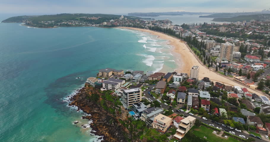 Aerial view vibrant coastline of Manly Beach in Sydney, featuring golden sand, turquoise ocean waves, bustling cityscape along the promenade, and surrounding residential suburbs. Drone flight panorama