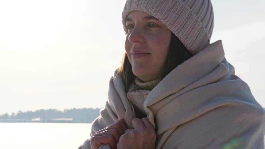 A woman stands by the water with her hands pressed together in a mindful pose. She wears a warm scarf and hat on a clear winter morning by the lake