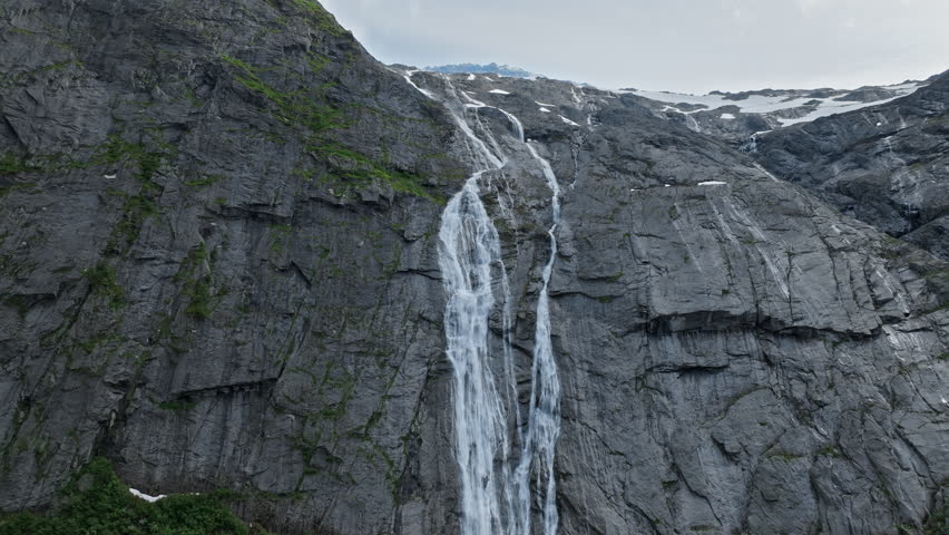 Tall waterfall cascading down rock wall near Briksdalsbreen in Norway. Aerial view captures flowing water, steep cliffs and lush greenery during summer.