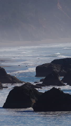 Large sea stacks rise from sandy beach on Oregon Coast as ocean waves roll in, with small human figures adding scale to dramatic Pacific shoreline landscape.