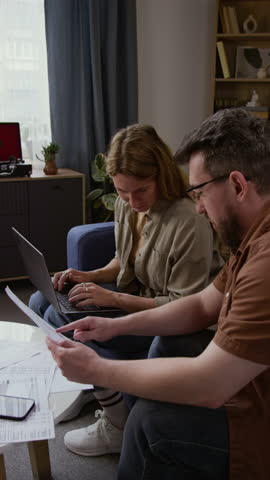 Vertical shot of Caucasian adult couple working with financial documents and using laptop while sitting on couch together