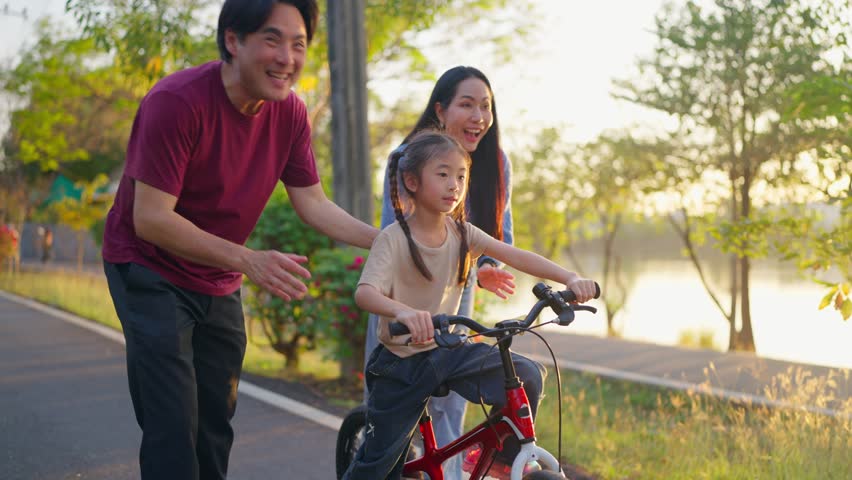 Asian young girl riding bicycle in garden while her parents look after. Adorable child feel happy and relax while learn to ride with family support, enjoying outdoor exercise activity in a public park