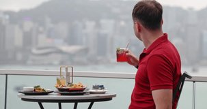 Man in red polo shirt sitting at rooftop bar holding cocktail, looking at Hong Kong skyline across Victoria Harbour. luxury travel, urban lifestyle, rooftop dining, vacation, city break, leisure, tour - Powered by Shutterstock - Get 15% off with code: PIKWIZARD15
