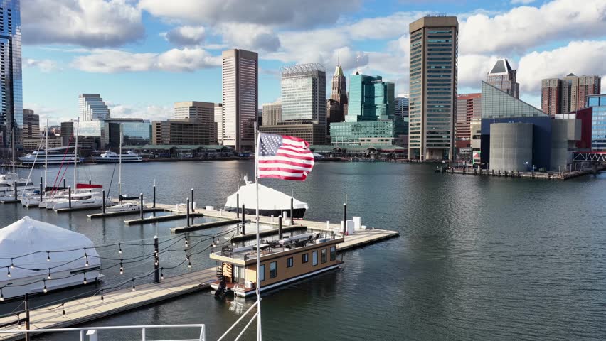 Aerial view of Baltimore’s Inner Harbor with an American flag in foreground. Marina docks, calm harbor waters and downtown skyline under bright daytime sky. Wide shot.