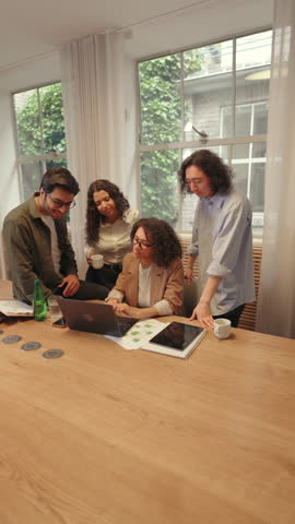 Diverse business people lean over a laptop in a bright office, analyzing project details and sharing insights, highlighting cooperation, problem solving, and a collaborative work culture.