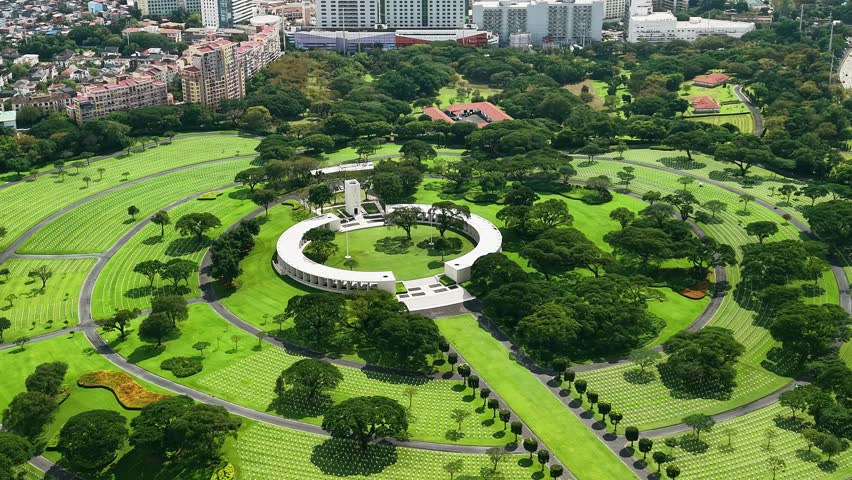 Philippines, Manila, January 27, 2026. Drone aerial over Manila American Cemetery and Memorial in Taguig, revealing circular monument, patterned headstones, and lush lawns with Metro Manila buildings in the distance.