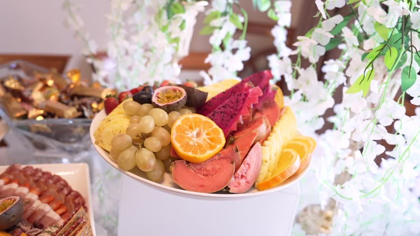 A collection of various fruits is set on a table during a gathering. The colorful selection includes sliced and whole fruits. Surrounding flowers add to the setup.