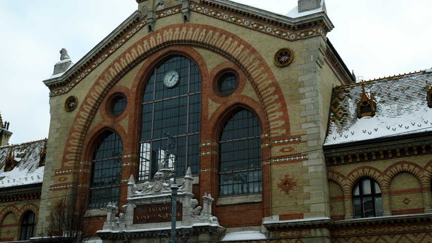 Exterior of the Great Market Hall in Budapest, Hungary, featuring historic brick architecture, arched windows, clock detail, and a winter atmosphere with light snow on the roof.
