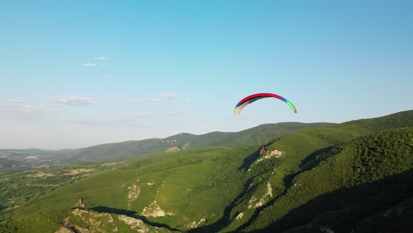 Drone aerial pan right to left of a paraglider flying above the green hills of Sokobanja, Serbia, with Ozren Mountain in the background. Scenic outdoor adventure view under a clear blue sky.