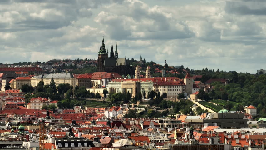 Telephoto aerial view of Prague Castle and St. Vitus Cathedral above red rooftops under dramatic clouds.