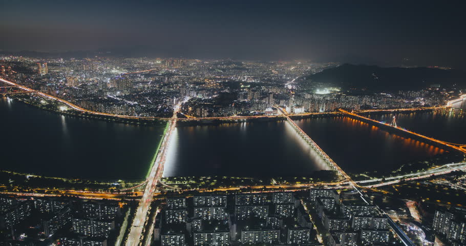 Aerial perspective captures a bustling city at night with bright lights reflecting on the water and bridges linking urban sections. Time-lapse, Seoul, South Korea
