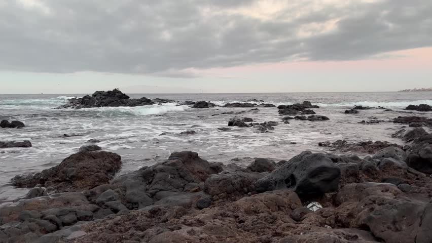 Waves washing over a pebble-covered shore in early daylight, ocean water covering and retreating across smooth stones along the rocky coastline