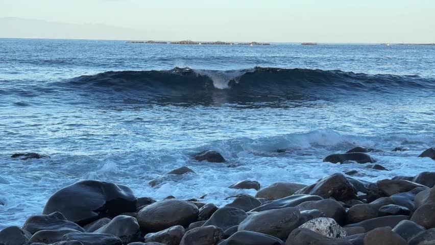 Waves washing over a pebble-covered shore in early daylight, ocean water covering and retreating across smooth stones along the rocky coastline