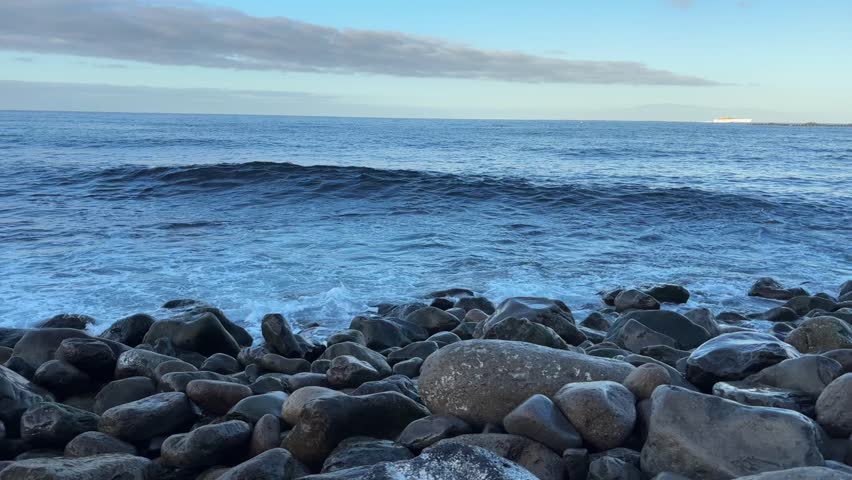 Waves washing over a pebble-covered shore in early daylight, ocean water covering and retreating across smooth stones along the rocky coastline