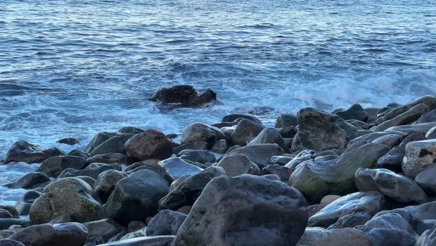 Waves washing over a pebble-covered shore in early daylight, ocean water covering and retreating across smooth stones along the rocky coastline