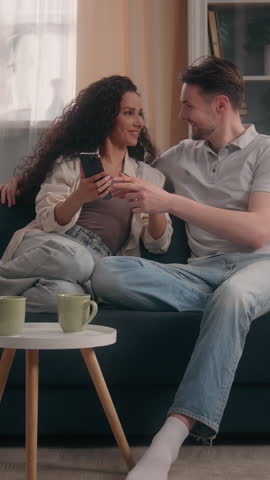 Multiethnic happy couple family at home using smartphone Caucasian man Hispanic Latina woman laughing mobile phone female male watch funny video shopping online smiling on couch enjoying moment indoor