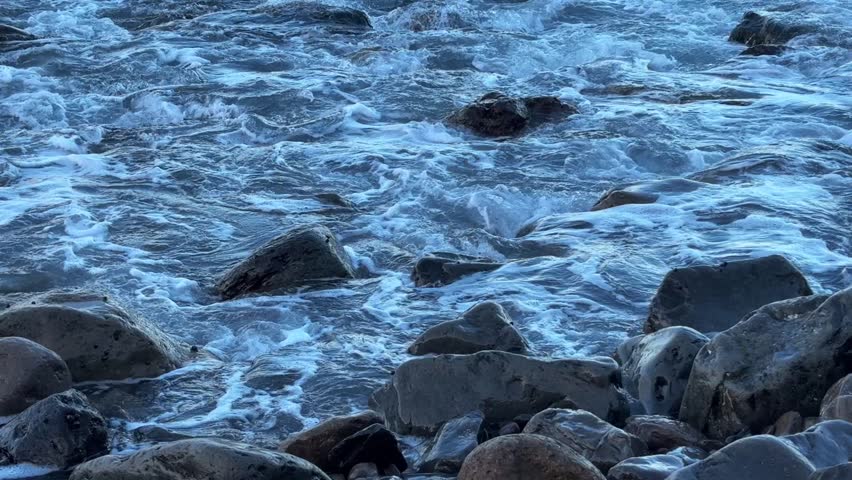 Waves advancing over rounded pebbles on a rocky shoreline at sunrise, ocean water moving steadily across the stones in soft early morning light