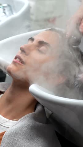 Relaxed woman with closed eyes receiving a therapeutic scalp massage and hair washing with steam at a professional hair salon, focusing on wellness and modern beauty care services