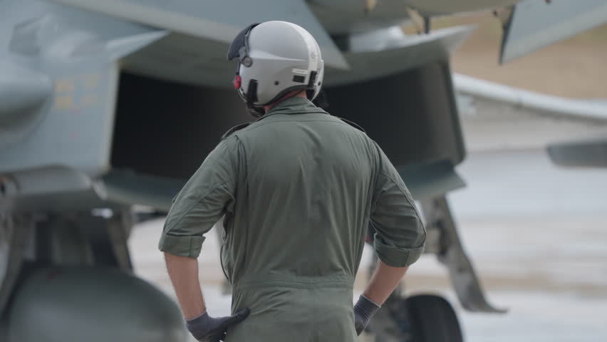 Rear view of a ground crew member in helmet and uniform standing near a fighter jet engine intake during preflight inspection on airbase apron