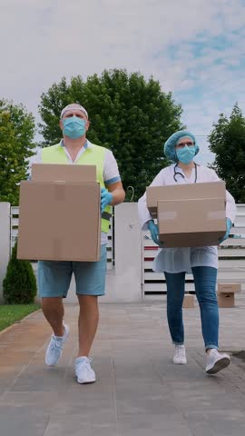Courier and doctor, in protective masks, gloves, going through courtyard of hospital or medical facility, carrying boxes with medical equipment, during coronavirus outbreak, quarantine.