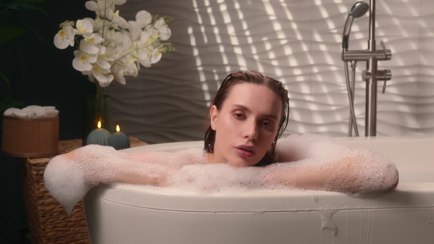 Caucasian woman relaxing in spa bathtub with candles diving foamy water female girl looking at camera enjoying calming home hotel bathroom atmosphere beauty bathing routine soothing wellness moment